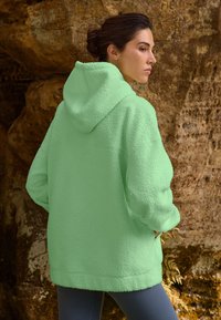 Green fleece hoodie with a textured surface, oversized fit, and a drawstring hood, paired with fitted gray pants.