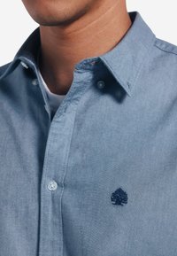 Camicia azzurra chiara in denim. Presenta un piccolo logo di un albero ricamato sul petto sinistro e un colletto di stile standard.