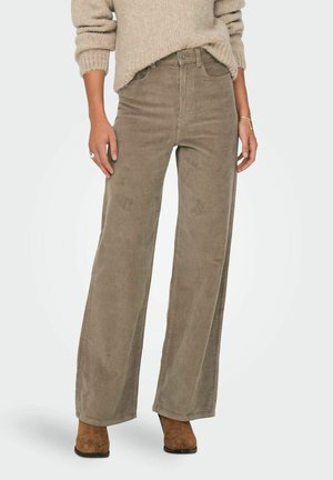 Kvinde iført beige bredbenede bukser i kordfløjl, en beige striktrøje, brune ruskindsstøvler til ankler og smykker med ring og armbånd.