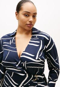 Σκούρο μπλε φόρεμα τύπου wrap με λευκές αφηρημέ νες γραμμές, που διαθέτει γιακά και χρυσή αγκράφα στη μέση, κατασκευασμένο από απαλό ύφασμα.