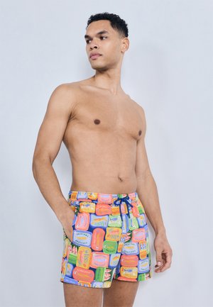 Homme torse nu aux cheveux courts et bouclés portant un short de bain coloré imprimé de divers designs de boîtes de sardines, debout devant un fond clair uni.