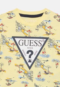 Κίτρινο βαμβακερό μπλουζάκι με εικονογράφηση παραθαλάσσιας σκηνής, μαύρο τριγωνικό λογότυπο με κείμενο και διακοσμητικά κουμπιά στον ώμο.