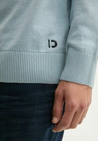 Lichtblauwe trui van zacht materiaal, met ribgebreide boorden en een zwart "C"-logo aan de onderkant van de linkerzijde. Close-up van de mouw en hand.