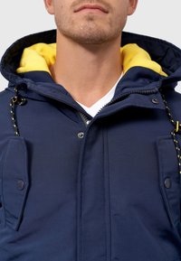 Veste à capuche bleu marine avec une doublure intérieure jaune, dotée d'une fermeture éclair, de boutons-pression et de deux poches poitrine. Tissu texturé avec des cordons réglables.