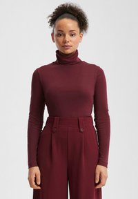 Bordeauxroter Rollkragenpullover aus weichem Stoff, figurbetontes Design. Trägt man zu hochtaillierten, weiten Hosen mit zwei Knöpfen vorne.