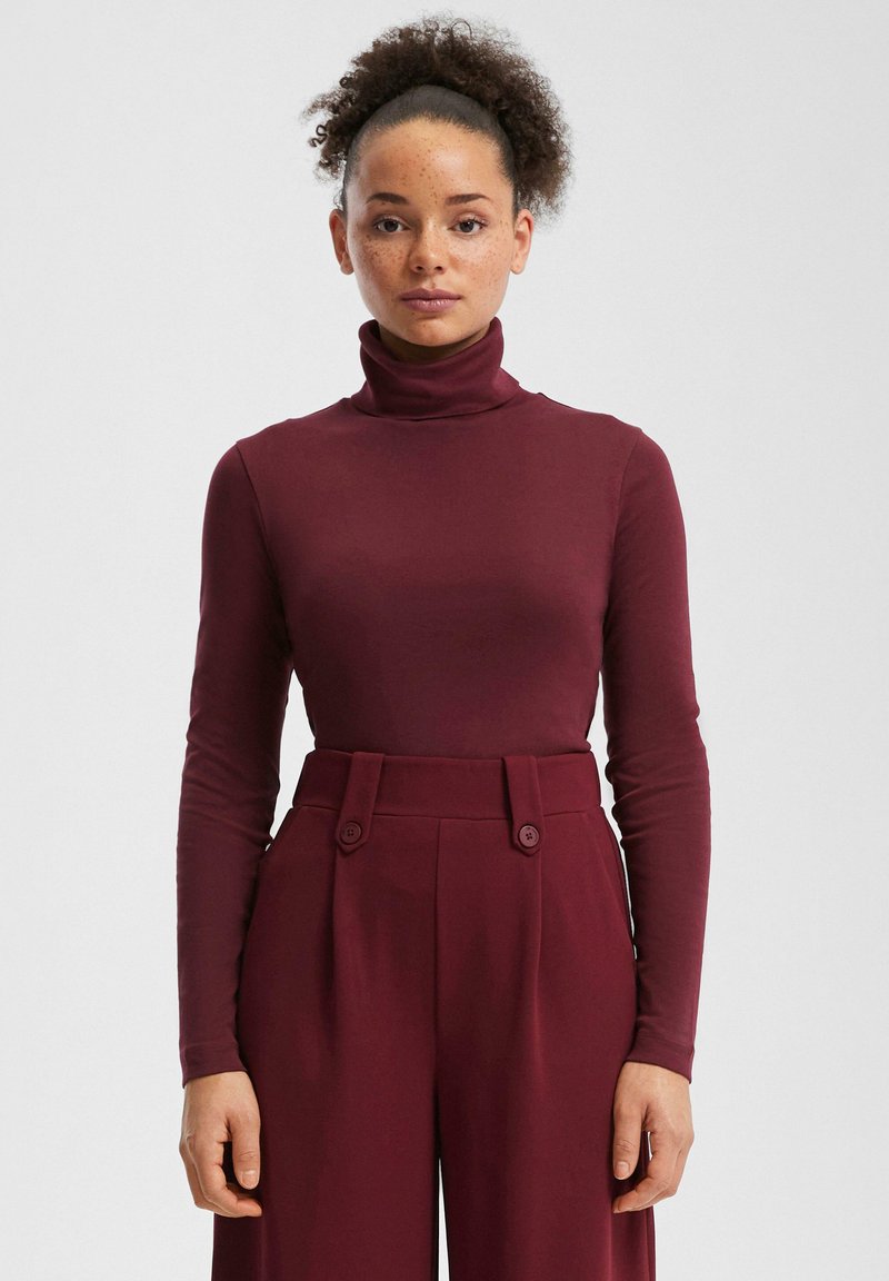 Bordeauxroter Rollkragenpullover aus weichem Stoff, figurbetontes Design. Trägt man zu hochtaillierten, weiten Hosen mit zwei Knöpfen vorne.
