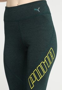 Leggings sportivi verde scuro con vita alta, tessuto strutturato e un audace logo giallo "PUMA" sulla coscia sinistra.
