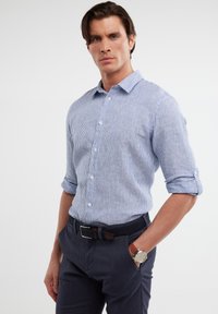 Camicia azzurra e bianca a righe con colletto aperto, maniche arrotolate e un aspetto tessile, abbinata a pantaloni scuri e una cintura.