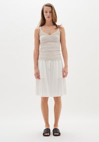 Helles beige Trägertop mit dünnen Trägern, kombiniert mit einem weißen knielangen Rock mit Kordelzug. Schwarze Rutschen-Sandalen runden das Outfit ab.