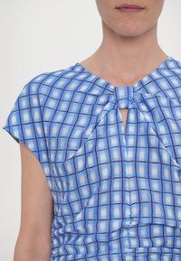 Femme portant un haut sans manches à carreaux bleus et blancs avec un petit pli frontal et un col arrondi, sur un fond uni.