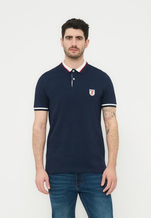 Άνδρας που φοράει μπλε ναυτικό πόλο με λευκή και κόκκινη επένδυση στο γιακά, μικρό έμβλημα στο στήθος και μπλε τζιν, στέκεται μπροστά σε απλό φόντο.