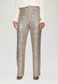 Hooggesneden, rechte broek bedekt met metallic gouden pailletten, gecombineerd met beige hakken en een vloeiende witte blouse met gerimpelde mouwen.