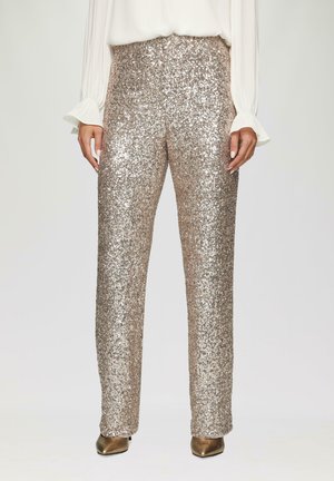 Hooggesneden, rechte broek bedekt met metallic gouden pailletten, gecombineerd met beige hakken en een vloeiende witte blouse met gerimpelde mouwen.