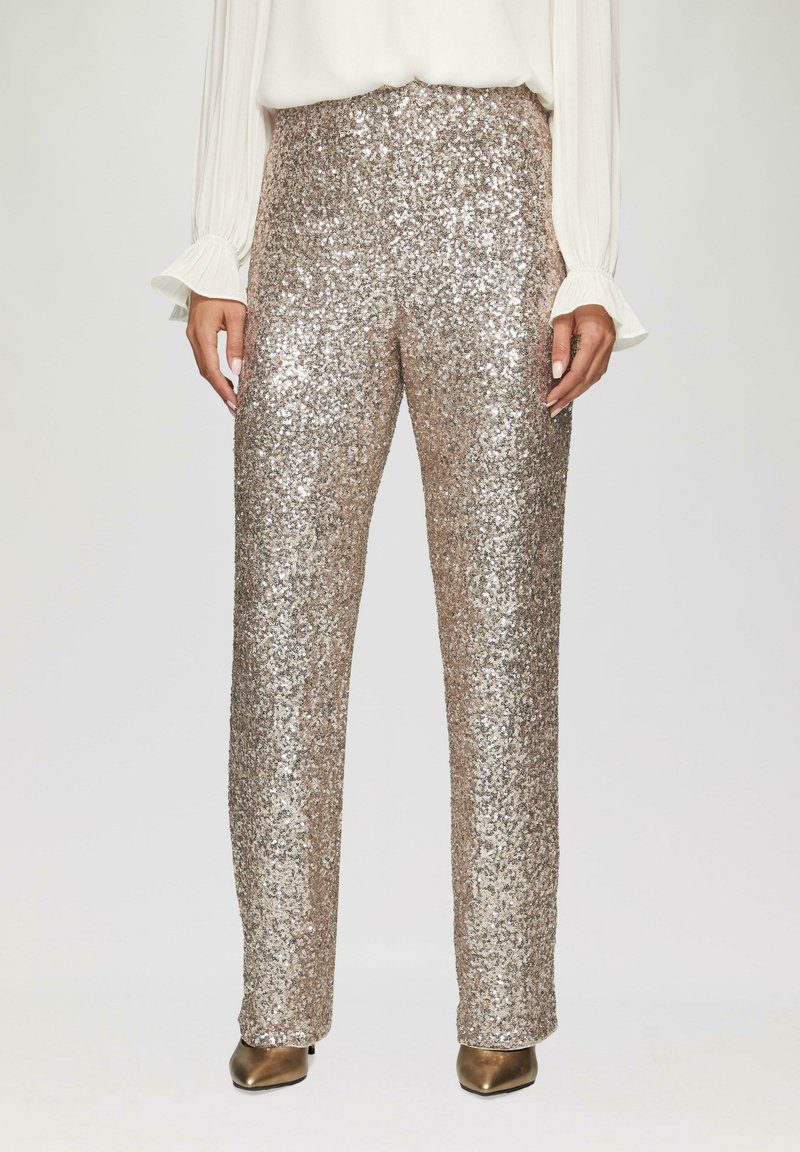 Hooggesneden, rechte broek bedekt met metallic gouden pailletten, gecombineerd met beige hakken en een vloeiende witte blouse met gerimpelde mouwen.