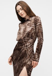 Γυναίκα που φοράει καφέ μακρυμάνικο φόρεμα με σχέδιο δέρματος ερπετού και πλευρική σχισμή, ποζάροντας μπροστά σε απλό λευκό φόντο.