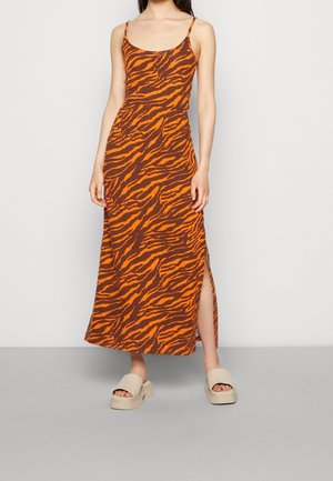 Kvinde iført en ærmeløs orange og brun tigerstribet maxikjole med sideslids og beige åbne platformssandaler.