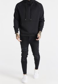 Felpa nera con zip e maniche lunghe, dotata di una tasca frontale e di un cordoncino. Indossata con jeans skinny neri strappati e sneakers bianche.