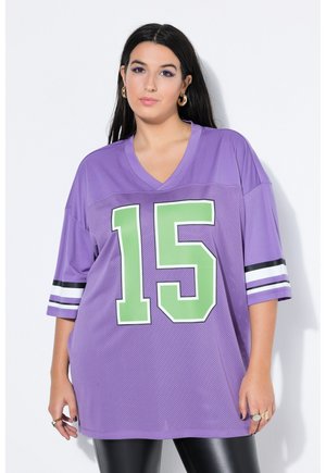 Jonge vrouw draagt een oversized paarse sporttrui met groen nummer 15, zwart-witte mouwstrepen en zwarte legging, staand tegen een witte muur.