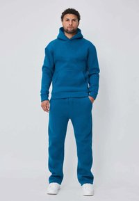 Sweat à capuche turquoise avec pantalon assorti en tissu doux. Le sweat à capuche a une poche kangourou ; les deux pièces ont des poignets et un ourlet côtelés. Baskets blanches.