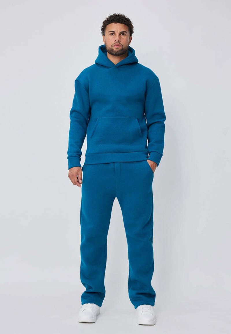 Sweat à capuche turquoise avec pantalon assorti en tissu doux. Le sweat à capuche a une poche kangourou ; les deux pièces ont des poignets et un ourlet côtelés. Baskets blanches.