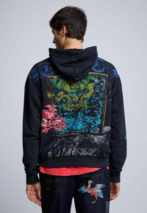 Man met zijn rug naar de kameraad, draagt een zwarte hoodie met kleurrijke draak- en bloemdessin en donkere spijkerbroek met geborduurde vogel op het achterzakje.