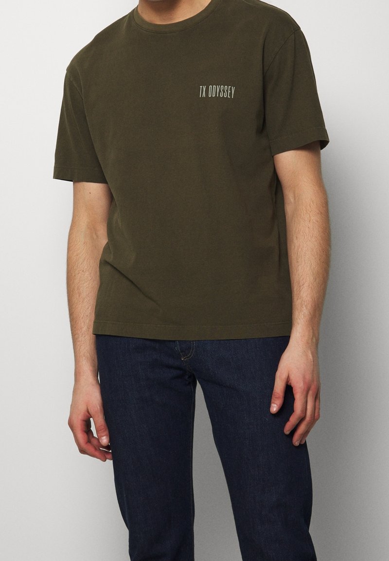 T-shirt en coton vert olive à manches courtes, col rond, avec un petit texte blanc « TX ODYSSEY » sur la poitrine. Texture lisse, coupe décontractée.