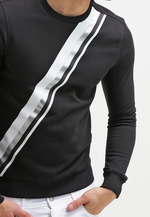 Czarna koszula z długim rękawem z ukośnymi metalicznymi srebrno-czarnymi paskami biegnącymi od ramienia do talii, noszona z białymi spodniami.