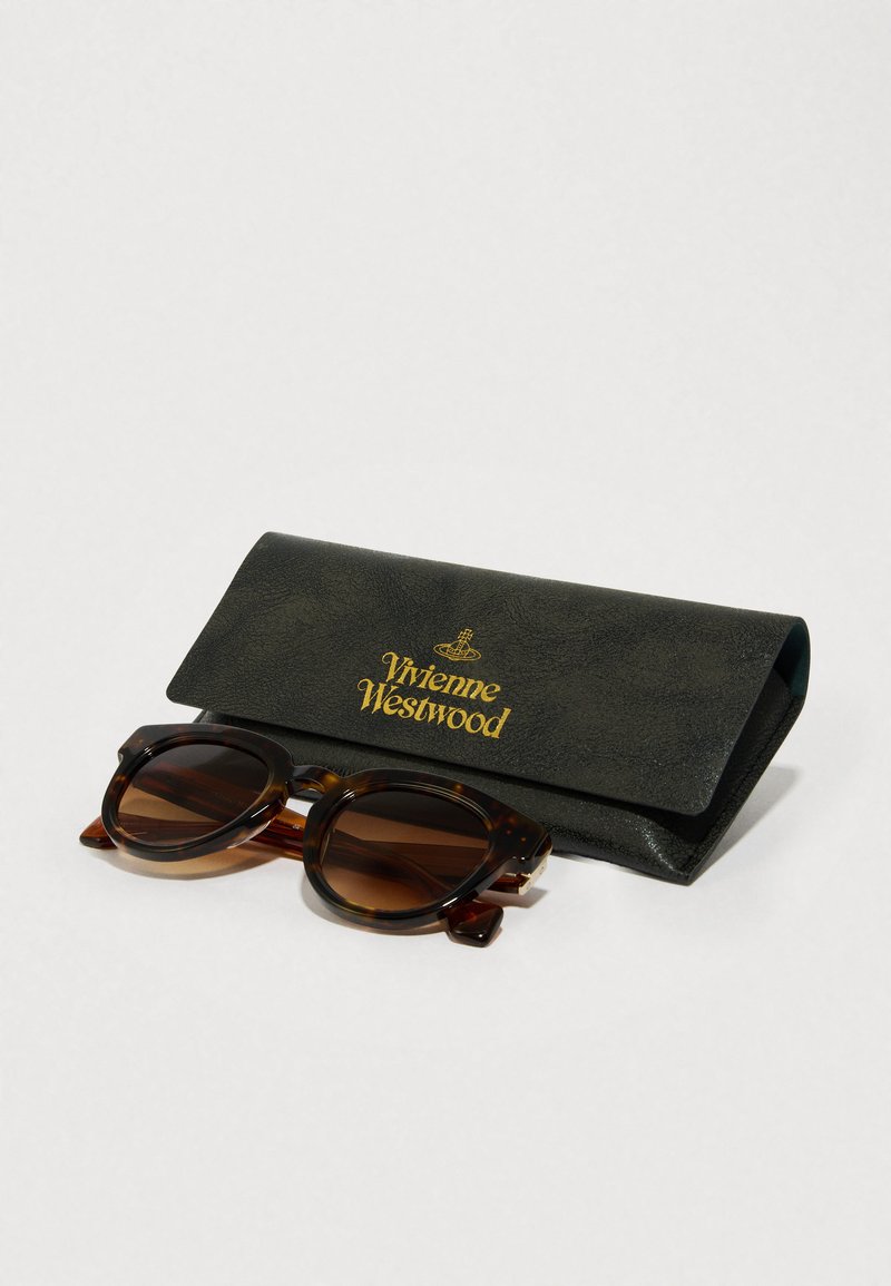 Brune skjoldere solbriller med runde rammer placeret ved siden af et sort læderetui prydet med guld "Vivienne Westwood" tekst.