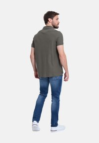 Polo shirt verde oliva de manga corta y con aberturas laterales, combinado con jeans azules y zapatillas blancas. Mostrado desde un ángulo trasero.