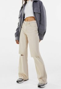 Beige wide-leg jeans with distressed sections, paired with a gray oversized jacket and a white cropped sweater. Subtle textures and colors.