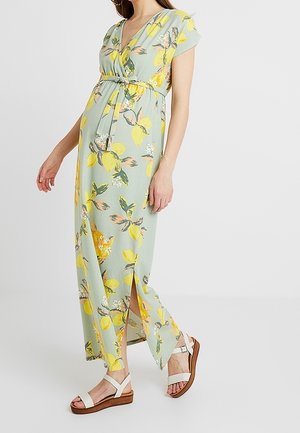 Femme portant une robe longue vert clair avec imprimé citrons jaunes et ceinture à nouer, associée à des sandales blanches, debout de profil.