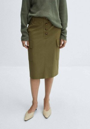 Vrouw draagt een olijfgroene kokerrok met knopen aan de voorkant en zakken, een groene gestructureerde trui met lange mouwen en beige puntige schoenen.
