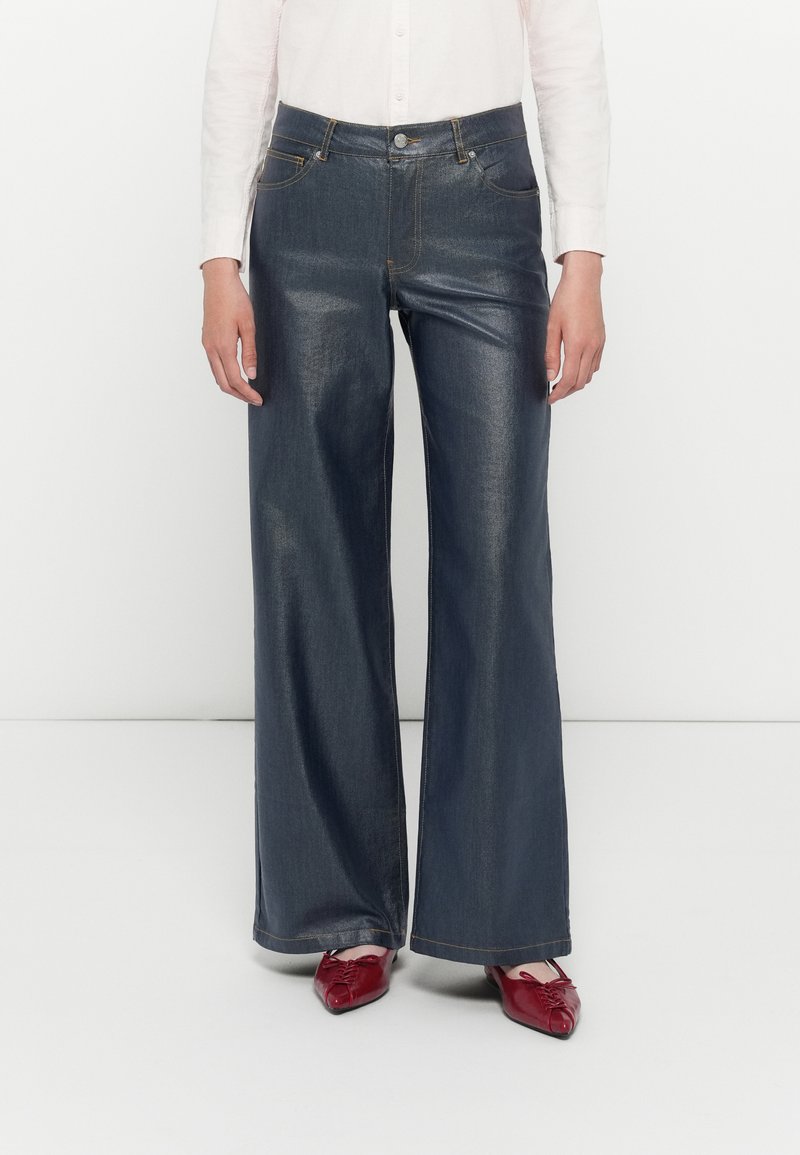 Pantalons larges en denim foncé avec taille haute. Comprend des poches traditionnelles et des coutures contrastées. Associés à des chaussures rouges.