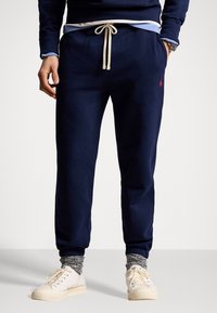 Pantaloni della tuta blu navy con vestibilità comoda, vita con coulisse e tasche laterali. Presentano un logo rosso sul fianco sinistro. Tessuto morbido.