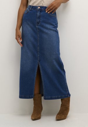 Femme portant une jupe midi en denim bleu avec une fente à l'avant et des bottines marron en suède sur fond blanc uni.