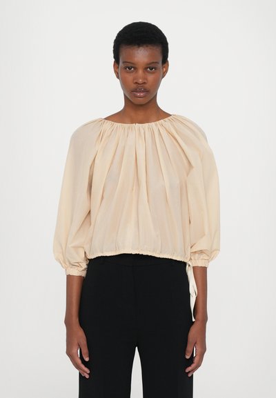 Blusa de color durazno pálido con mangas abullonadas, escote fruncido y cinturilla elástica. Tejido ligero con una textura suave y costuras mínimas visibles.