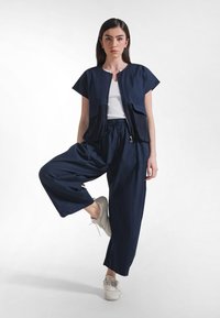 Granatowa krótka kurtka z krótkimi rękawami, z zamkami błyskawicznymi, w zestawie z luźnymi granatowymi spodniami. Model ma na sobie białą koszulkę i jasnoszare tenisówki.