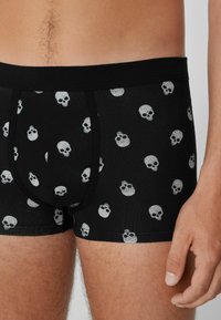 Černé boxerky se vzorem malých bílých lebek, které má na sobě muž, viditelné dolní torso, boky a pravá ruka.