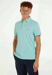 Jovem com cabelo ruivo encaracolado e sardas, vestindo uma camisa polo azul clara e calças escuras, de pé com uma mão no bolso.