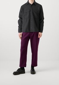 Czarna tkana koszulowa kurtka z dwoma kieszeniami na piersi, zestawiona z luźnymi spodniami w głębokim fioletowym kolorze oraz czarnymi sznurowanymi butami na grubej podeszwie.