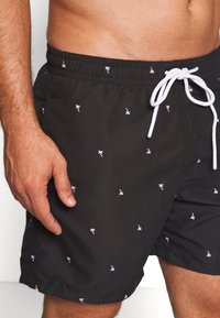 Fekete fürdőshorts fehér pálmafa mintával, rugalmas derékpánttal és fehér húzózsinórral. Az anyag könnyűnek és simának tűnik.