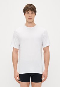 Balta trumpa rankovėmis marškinėliai, pagaminti iš lygaus medvilnės audinio, su klasikine apykakle ir tiesia apačia. Modelis vilki juodas šortus.