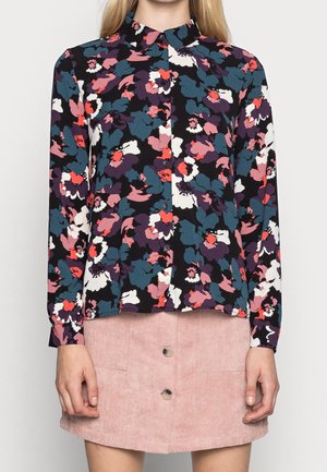 Vrouw draagt een zwarte blouse met lange mouwen met een meerkleurige bloemenprint en een lichtroze corduroy rokje met knopen.