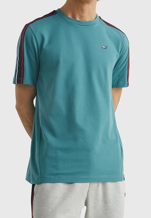 Mand iført en teal kortærmet t-shirt med røde og sorte striber på ærmerne og et lille logo på brystet, kombineret med lysegrå shorts.