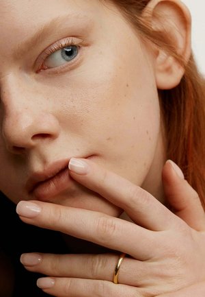 Primer plano de un rostro con piel clara, ojo azul, textura suave y ligeras pecas. Dedos con uñas en tono nude tocan los labios. Anillo de oro en uno de los dedos.