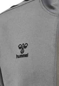 Sweatshirt gris avec une fermeture éclair et un logo noir sur la poitrine gauche. Le tissu a une texture lisse avec une coupe décontractée.