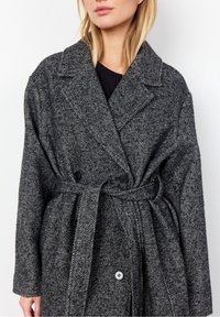 Szary płaszcz w jodełkę z podwójnym zapięciem, dużymi klapami, paskiem w talii oraz dwoma czarnymi guzikami z przodu.