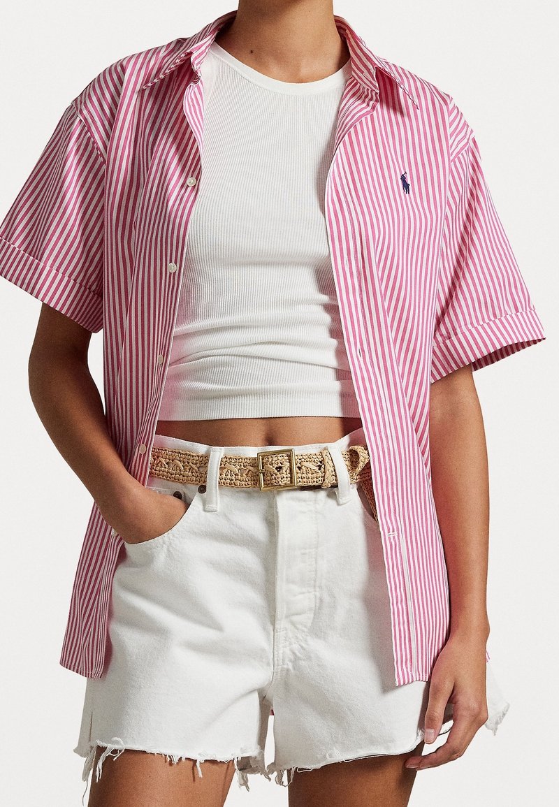 Camicetta a maniche corte con bottoni in rosa e bianco a righe, caratterizzata da un colletto classico. Abbinata a pantaloni corti bianchi logorati e una cintura intrecciata.