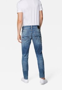 Jeans de denim azules para hombre con un ajuste slim, que presentan secciones descoloridas, dos bolsillos traseros y una etiqueta de cuero en la cinturilla. Llevados con una camiseta blanca.