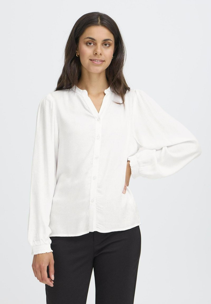 Camicia bianca con colletto, chiusura a bottoni e maniche lunghe a sbuffo. Realizzata in un leggero tessuto testurizzato con polsini arricciati. Abbinata a pantaloni neri.